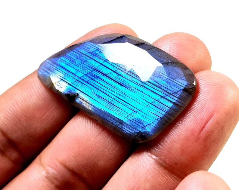 May include: A blue and iridescent labradorite gemstone with a faceted, rectangular shape. The gemstone is held in a hand.