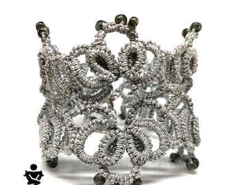 Puede incluir: Un brazalete de crochet plateado con cuentas de color gris oscuro. El brazalete está hecho de un delicado y complejo patrón de bucles y nudos.