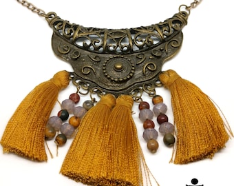 May include: A gold necklace with a large, ornate pendant and multiple yellow tassels. The pendant is a crescent shape with intricate designs and a small circle in the center. The tassels are made of thin strands of yarn and are attached to the pendant with small beads.