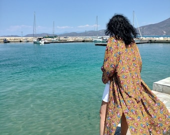 May include: A woman wearing a long, yellow and pink floral print kimono with a white skirt, standing on a stone pier overlooking a body of water. The water is a vibrant turquoise blue and there are boats in the distance. The sky is a clear blue.