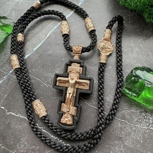 Könnte beinhalten: Eine schwarze geflochtene Kordelkette mit einem goldfarbenen Kreuzanhänger. Das Kreuz zeigt eine detaillierte Darstellung von Jesus Christus. Goldfarbene zylindrische Perlen sind entlang der Kordel angeordnet.
