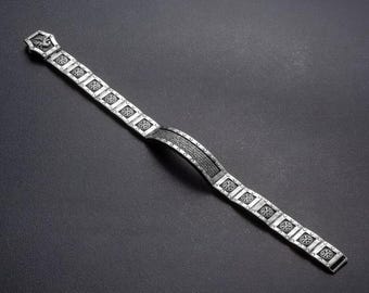 May include: A silver-toned bracelet with an ornate design. The bracelet features a series of square links with a floral pattern, connected by a curved central section. The clasp has a decorative element. The bracelet is displayed on a dark surface.