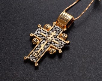 Może przedstawiać: Złoty i srebrny wisiorek w kształcie krzyża. Wisiorek posiada skomplikowane wzory celtyckie i ikonografię religijną. Krzyż wisi na cienkim złotym łańcuszku na ciemnym tle.