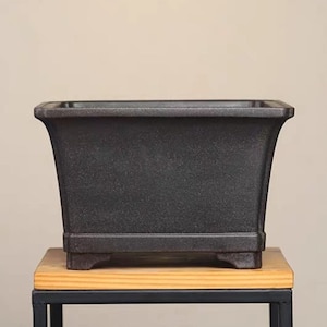 Pode incluir: Um vaso quadrado de cerâmica preto com uma superfície texturizada e quatro pequenos pés. O vaso está sentado em uma mesa de madeira com uma moldura de metal preto.