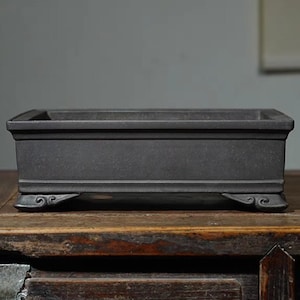 Może przedstawiać: Prostokątna, czarna ceramiczna doniczka do bonsai z podwyższonym rantem i ozdobnymi nóżkami. Doniczka stoi na drewnianej powierzchni.