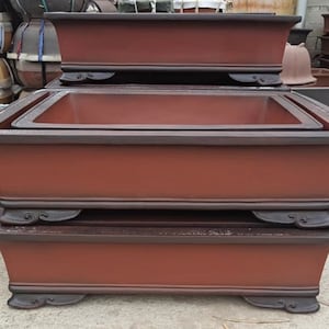 Puede incluir: Cuatro macetas rectangulares de bonsai de terracota apiladas con bordes marrones oscuros y pies decorativos. Las macetas están dispuestas en forma de pirámide.