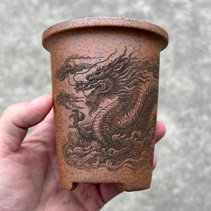 May include: A terracotta-coloured ceramic pot with a detailed black dragon design. The pot has a rounded base and a wide opening.