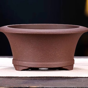 Può includere: Un vaso per bonsai in ceramica marrone con forma arrotondata e tre piccoli piedini. Il vaso ha una superficie ruvida e testurizzata.