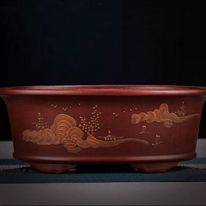 Puede incluir: Una maceta de bonsái de cerámica ovalada roja con un diseño de paisaje en pintura dorada. La maceta tiene tres pequeños pies.