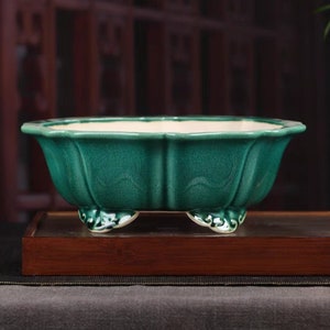 May include: A green ceramic planter with a rounded, scalloped rim and four decorative feet. The planter is designed to hold plants and flowers.