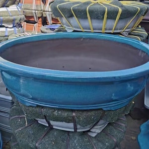 Può includere: Un grande vaso ovale in ceramica di colore blu-verde con interno marrone scuro. Il vaso ha un bordo leggermente rialzato e una finitura liscia e lucida.