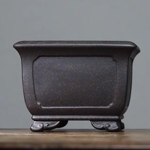 Puede incluir: Una maceta cuadrada de cerámica negra con una superficie texturizada y cuatro patas decorativas.