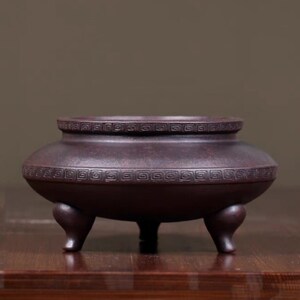 May include: A dark brown ceramic incense burner with three short legs. The round body features a decorative geometric pattern around the rim and midsection. The incense burner is designed to hold incense.