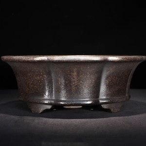 May include: A dark brown ceramic bonsai pot with a rounded shape and a flared rim. The pot has a textured surface and rests on three small feet.