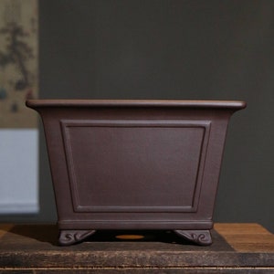 May include: A dark brown square ceramic bonsai pot with a decorative rim and four small feet.