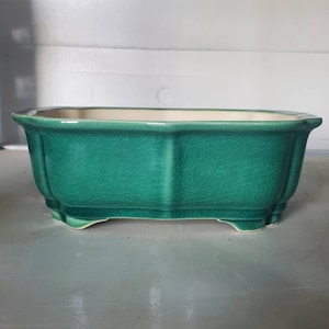 Può includere: Un vaso da fiori in ceramica verde con forma rettangolare e quattro piccoli piedini. Il vaso ha una finitura smaltata craquelé.