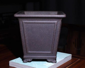 Puede incluir: Una maceta cuadrada marrón oscuro con una superficie texturizada. La maceta tiene un diseño clásico con paneles decorativos y descansa sobre pequeños pies ornamentados. Está sobre una pila de papel azul claro.