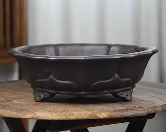 May include: A dark brown ceramic planter with a scalloped rim and three feet. The planter is sitting on a wooden table.