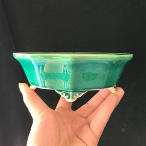 Puede incluir: Un macetero hexagonal de cerámica verde con tres patas. El macetero tiene un acabado brillante y un esmalte verde claro.