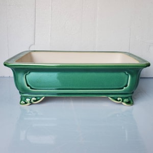 May include: A green ceramic bonsai pot with a white glazed interior. The pot has a rectangular shape with rounded corners and decorative details on the sides. It has three feet on the bottom.