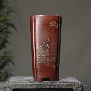 May include: A terracotta-coloured ceramic planter with a rectangular shape and a decorative design of a mountain scene with clouds and birds. The planter has drainage holes and a slightly flared rim.