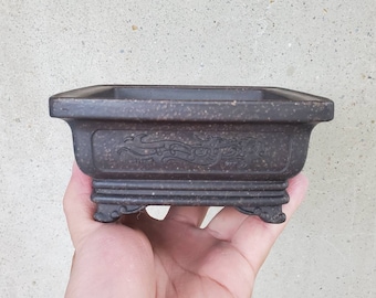 May include: A square, dark brown bonsai pot with a raised rim and three legs. The pot has a decorative design of a dragon and waves.
