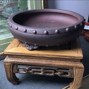 Könnte beinhalten: Ein dunkelbrauner Keramik-Bonsai-Topf mit einer strukturierten Oberfläche und einem erhöhten Rand mit kleinen, runden Akzenten. Der Topf steht auf einem Holzständer mit aufwendigen Schnitzereien.