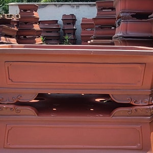 Może przedstawiać: Kolekcja prostokątnych donic do bonsai w kolorze terakoty. Donice mają dekoracyjną podstawę ze zdobieniami i prostokątny wierzch z wpuszczonym panelem. Donice są w różnych rozmiarach i kształtach, odpowiednie do bonsai.