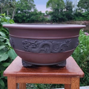 May include: A dark brown, round ceramic planter featuring a wide, flat rim and a raised, sculpted dragon relief pattern encircling the exterior. The vessel is supported by three small, integrated feet at the base, showcasing a matte finish.