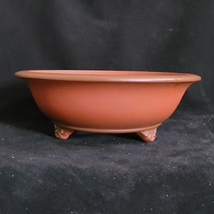 以下が含まれることがあります： 丸みを帯びた形をした、3つの小さな足が付いたテラコッタ製の盆栽鉢。鉢は赤褐色です。