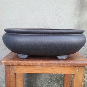 May include: A dark gray, oval-shaped bonsai pot with a textured surface. The pot has a wide, shallow design and rests on three small feet. The pot is sitting on a small wooden table.