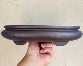 May include: A dark brown, oval-shaped bonsai pot with a rounded base and three small feet. The pot is held in a hand, showcasing its smooth, textured surface. The pot is designed for planting small trees.