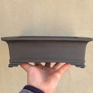 以下が含まれることがあります： テクスチャ加工された表面とわずかに盛り上がった縁を持つ、濃い茶色の長方形の盆栽鉢。鉢には小さな足が4つあります。