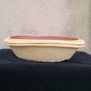 May include: A handmade, oval-shaped ceramic planter with a light beige exterior and a reddish-brown interior. The planter has a layered rim design and sits on a black surface. The overall aesthetic is rustic and earthy.