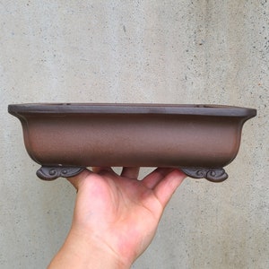 Puede incluir: Un macetero rectangular de cerámica marrón con un fondo ligeramente redondeado y dos pies decorativos a cada lado. El macetero está hecho de terracota sin esmaltar.