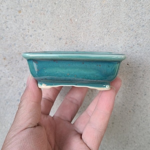 Op de afbeelding: Een kleine, rechthoekige, teal-kleurige keramische bonsai pot met een licht verhoogde rand en een getextureerde glazuur. De pot heeft vier kleine voetjes aan de onderkant.