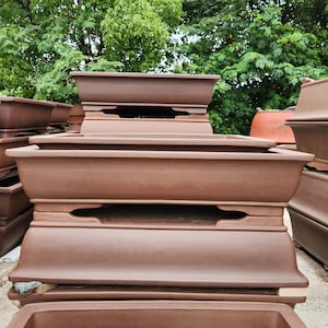 Puede incluir: Una colección de macetas de bonsái marrones rectangulares apiladas. Las macetas tienen un diseño clásico con un borde ligeramente ensanchado y cortes decorativos en los lados. Las macetas están hechas de un material similar a la terracota y están dispuestas al aire libre.