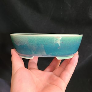 Könnte beinhalten: Ein ovaler Keramik-Pflanztopf in Hellblau und Grün mit weißem Rand und drei kleinen Füßen. Der Topf ist aus glasierter Keramik und hat eine gesprenkelte Oberfläche.