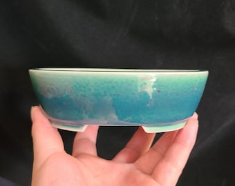May include: A light blue and green oval ceramic planter with a white rim and three small feet. The planter is made of glazed pottery and has a speckled finish.