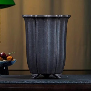 Pode incluir: Um vaso de cerâmica cinza escuro com um design canelado e três pequenos pés. O vaso está sobre uma superfície de ardósia preta.