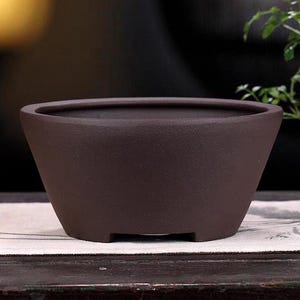 May include: A dark brown ceramic bonsai pot with a rounded oval shape and a slightly flared rim. The pot has a textured surface and three small feet on the bottom.