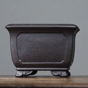 Puede incluir: Un macetero cuadrado de cerámica marrón oscuro con una superficie texturizada y cuatro patas decorativas. El macetero tiene un borde ligeramente acampanado y una forma rectangular.