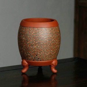 Puede incluir: Una tetera de arcilla roja con un diseño estampado en marrón y verde. La tetera tiene tres patas y un cuerpo redondo.