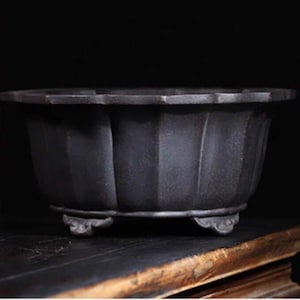 Puede incluir: Una maceta de cerámica negra con un diseño acanalado y tres patas. La maceta está sobre una superficie de madera.