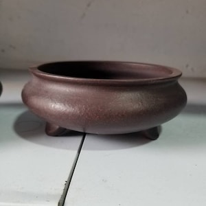 Puede incluir: Una maceta de bonsái de cerámica marrón oscuro con tres patas. La maceta tiene una forma redondeada y una superficie lisa ligeramente texturizada.