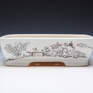 Può includere: Vaso rettangolare in ceramica bianca con una scena di paesaggio dipinta in nero e rosso. La scena include alberi, un ponte, una montagna e un piccolo edificio. Il vaso ha una base rialzata.