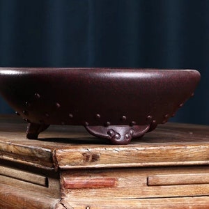 Peut inclure: Un pot en céramique rond de couleur rouge foncé avec trois pieds. Le pot a une surface texturée et est posé sur une table en bois.