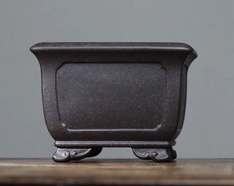 Puede incluir: Un macetero cuadrado de cerámica marrón oscuro con una superficie texturizada y cuatro patas decorativas. El macetero tiene un borde ligeramente acampanado y una forma rectangular.