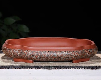 May include: A red clay bonsai pot with a decorative design. The pot has a rounded rectangular shape with four short legs.