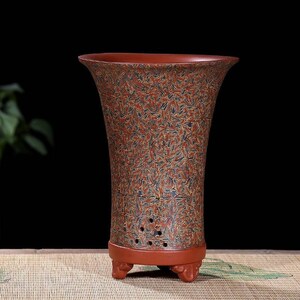 Puede incluir: Una maceta de arcilla roja con un patrón decorativo de líneas azules y doradas. La maceta tiene una abertura ancha y una base estrecha con tres patas.